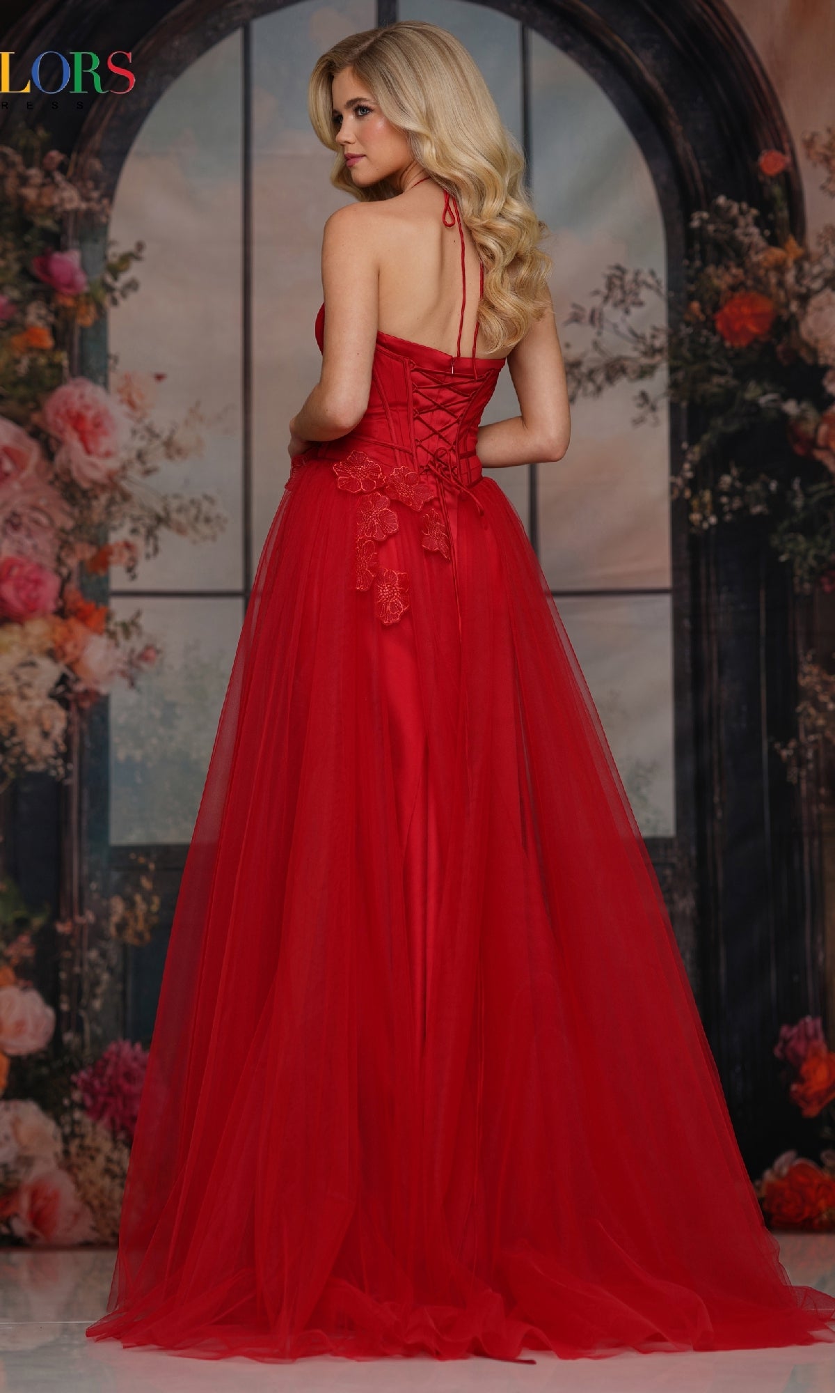 Wearing the Colors Dress 3796, a woman stands before a floral arched window backdrop. The long prom dress features a lace-up back, 3D floral appliques, and an elegant A-line tulle skirt as she looks over her shoulder.